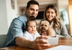 A family of two parents, one child, and a dog are smiling as they take a selfie together.