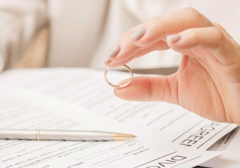 A hand holds a wedding ring over divorce paperwork, with a pen in the background.