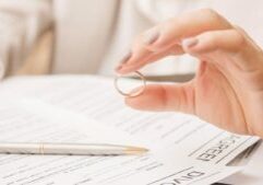 A hand holds a wedding ring over divorce paperwork, with a pen in the background.