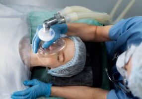 An anesthesiologist in a surgical gown and mask carefully administering anesthesia to a patient through a mask, while the patient lies on the operating table, preparing for surgery.