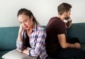 A couple sitting on opposite ends of a teal couch showing signs of relationship distress. A woman in a pink plaid shirt sits on the left looking downward with her hand on her face, appearing sad or worried. A man in a burgundy t-shirt sits on the right with his back partially turned, also with hand on face, suggesting tension or disagreement between them.