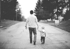 Father and young son walking hand in hand down a tree-lined country road, viewed from behind in black and white.