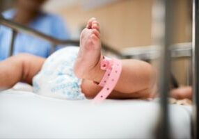 A newborn baby lying in a hospital bassinet with a pink identification bracelet around its ankle. The infant's legs are visible, and a healthcare professional is blurred in the background. The image relates to birth injuries, highlighting the vulnerability of newborns during the delivery process.