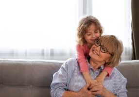 A smiling grandmother in a blue and white striped shirt and glasses embraces her young granddaughter, who is wearing a pink shirt. They are sitting together on a gray couch, sharing a warm, affectionate moment with the child hugging her grandmother from behind.