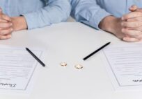 Two people sitting at a table with divorce papers, wedding rings, and pens, symbolizing the process of ending a marriage.