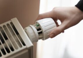 Close-up of a hand adjusting a white thermostatic valve on a home radiator with vertical slots. This heating equipment requires proper maintenance and carbon monoxide detectors to prevent CO poisoning, ensure safety compliance, and protect occupants' legal rights.