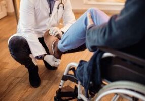 A doctor wearing gloves examines the leg of a senior patient in a wheelchair, assessing issues related to amputation and loss of limb function.