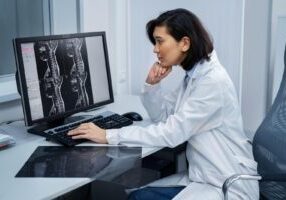 A doctor sits at her desk, studying a CT scan of a spinal cord displayed on her computer screen, focusing on detailed spinal analysis and diagnosis.