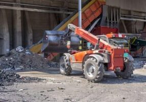 Construction trucks are positioned at a demolition site, with debris and dust surrounding them.