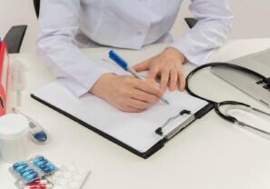 Close-up of a doctor's hand writing a prescription on paper, with a pen in hand. Pills are scattered on the desk, highlighting issues related to medication, prescriptions, and patient care.