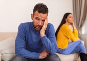 A distressed man sits on a couch, his face resting in his hand, while his partner turns away, highlighting the emotional toll of infidelity in marriage. When adultery becomes a factor in divorce, it can impact property division, alimony, and child custody.