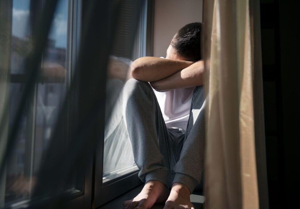 Person sitting on window sill, head buried in arms, conveying distress and isolation after traumatic experience.