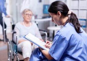 An image depicting a healthcare professional attentively taking notes while seated near an elderly woman in a wheelchair. This highlights the importance of vigilance in identifying signs of elder abuse in nursing homes, ensuring proper reporting, and taking legal action to protect vulnerable individuals.