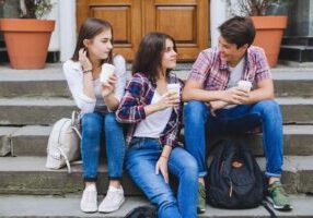 Three teenagers sitting on outdoor steps in front of a wooden door. They're all holding coffee cups and wearing casual clothes with plaid shirts and jeans.
