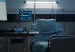 An empty hospital bed in a dimly lit room, with medical monitoring equipment displaying vital signs on a screen.