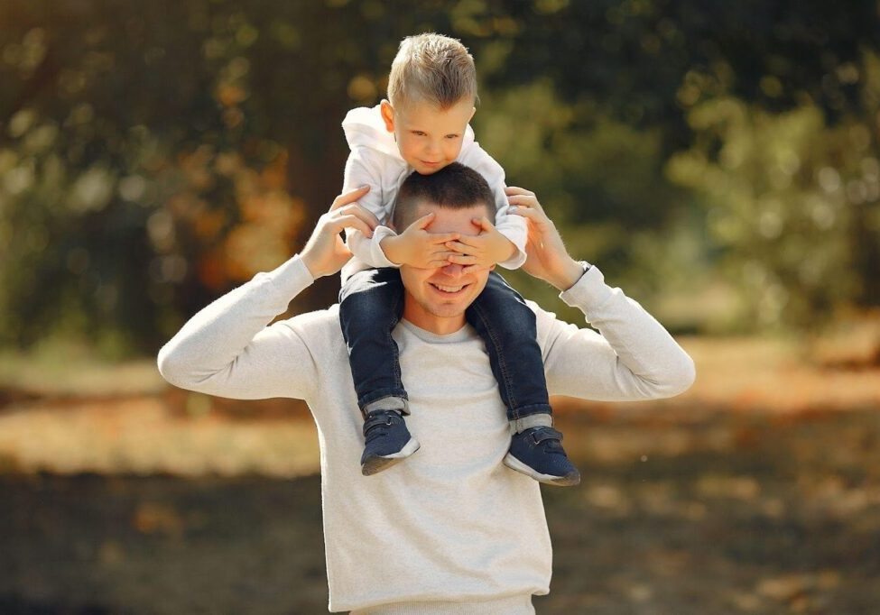 A father carrying his child on his shoulders, both smiling and enjoying an outdoor moment. The image conveys the topic of FAQs about paternity in Alabama, focusing on fatherhood and parental rights.