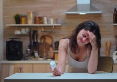 A woman sitting at a table holding pills, appearing unwell, symbolizing the effects of foodborne illness. This image relates to foodborne illness and contamination, emphasizing issues of liability and the potential for compensation in cases of negligence.