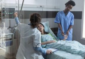 Two medical professionals attending to a patient in a hospital bed: one nurse placing a pulse oximeter on the patient's finger, while a doctor adjusts an IV drip.