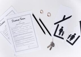 Flat lay of divorce paperwork including divorce forms, two wedding rings, house keys, and family pictogram symbols suggesting separation or division of family unit. The image includes black pens and is set against a white background.