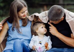 A young family sits together under a blanket, with parents smiling at their toddler who is wearing a light gray sweater and holding toys. The mother wears a blue denim sleeveless top, while the father wears a black shirt.