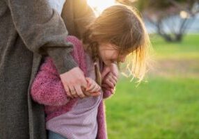 A young child with light blonde hair wearing a pink knit sweater and striped shirt stands close to an adult in a gray coat. The child is holding the adult's hand, which has a ring on it. They are outdoors in a park or grassy area with soft sunlight creating a warm backlit glow around the child's hair.