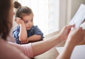 A young girl with her hair in a top knot sits at a table, resting her head on her hand and looking thoughtfully to the side. An adult's hands can be seen holding a document in the foreground, suggesting a serious or contemplative moment between a child and an adult.