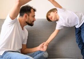 A father and son engage in a playful moment, with the father sitting on a gray couch and holding his son's hand. The son leans towards his father with a smile, while both have their other hands raised, suggesting a lighthearted interaction between parent and child.