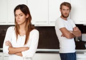 A couple standing apart in a kitchen, both with arms crossed. The woman stands in the foreground looking upset, while the man stands in the background appearing frustrated. They're both wearing white tops, suggesting tension or conflict in their relationship.