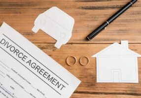 A divorce agreement document on a wooden table with paper cutouts of a house and car, two wedding rings, and a black pen, representing property division during divorce proceedings.