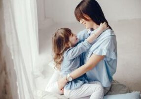 A woman with long dark hair in a light blue top sits by a window embracing a young girl with long light brown hair. Both are wearing matching blue tops and white pants, creating a warm, intimate mother-daughter moment in a bright, minimalist setting.
