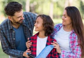 A family of three smiling together outdoors. A father in a gray plaid shirt and a mother in a pink plaid shirt stand on either side of their young daughter, who has curly hair and is wearing a red plaid shirt.