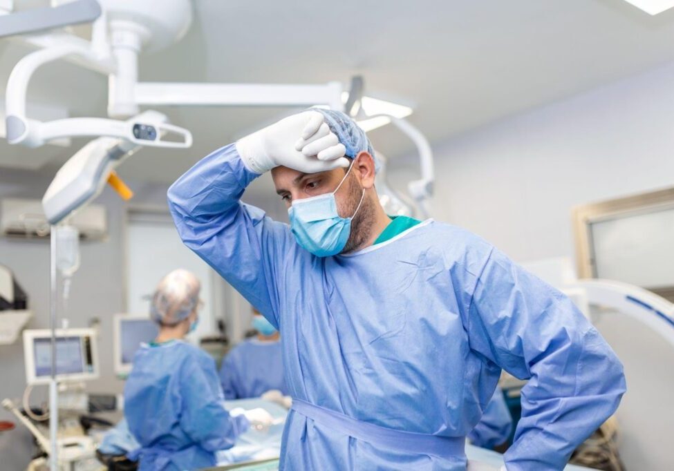 A distressed doctor stands in a surgery room, appearing deep in thought, with medical equipment in the background. The image conveys the high-stress environment of surgical procedures, underscoring the serious implications of surgical errors and the potential for medical malpractice.