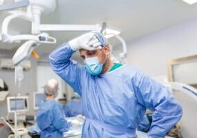 A distressed doctor stands in a surgery room, appearing deep in thought, with medical equipment in the background. The image conveys the high-stress environment of surgical procedures, underscoring the serious implications of surgical errors and the potential for medical malpractice.