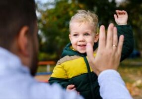 A smiling child raising his hand to high-five his dad, who adopted him, as they stand together outside. This joyful moment highlights the strong bond between them and reflects the journey of adoption, while also symbolizing the choice between open adoption and closed adoption.