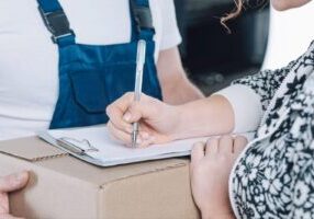 A woman signs documents related to product liability, with a box containing the product in front of her. The image represents the legal process involved in addressing issues of product responsibility and consumer safety.