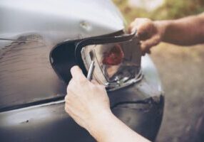 A hand carefully fixes the front light of a car that has been involved in a rear-end collision. The light appears damaged, with slight dents on the vehicle’s front, indicating the aftermath of the accident and the process of repairing the car.