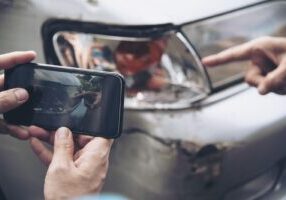 Hand holding smartphone, photographing crumpled rear bumper of car after a rear-end collision.