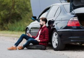 A frustrated driver sits on his phone beside his damaged car after a rear-end collision on an Alabama highway.