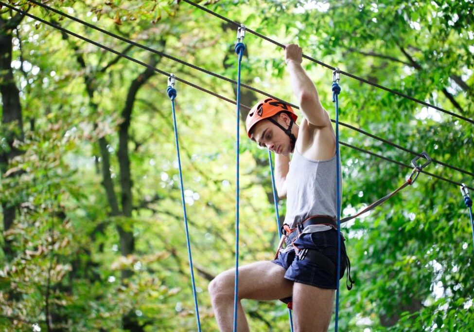 A person wearing a white tank top, navy shorts, orange climbing helmet, and safety harness navigates a high ropes course. They are gripping blue safety cables while moving between platforms, surrounded by green forest canopy.