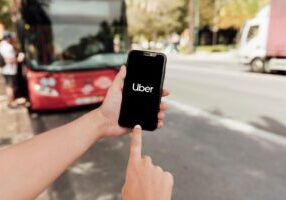 A person holding a smartphone with the Uber app open, standing near a city street with a bus and vehicles in the background. This image represents the context of rideshare driver liability, emphasizing the importance of understanding legal responsibilities and protections when using services like Uber, Lyft, and similar platforms.