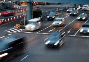 An aerial view of a busy intersection at dusk or night, showing cars in motion with blurred headlights and taillights indicating fast movement. The road is marked with traffic lanes and features orange safety cones and road barriers. The motion blur of vehicles suggests rapid traffic flow, while safety measures like road markings and barriers are clearly visible, highlighting the context of traffic safety and potential road incidents. The scene captures the dynamic and potentially dangerous nature of urban traffic conditions.