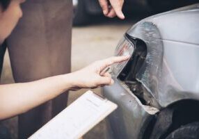 Insurance adjuster in a suit taking notes on a clipboard while examining a damaged vehicle at a T-bone accident scene.