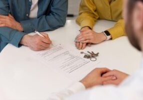 Two people sit at a white table, with one person in a blue shirt signing a document while another in a yellow shirt watches. A set of keys is placed on the document, suggesting a legal or contractual meeting, related to real estate.