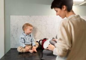 A young toddler with light brown hair wearing a blue button-up shirt and beige pants sits on a dark countertop against a marble wall. A person with short dark hair wearing a beige sweater stands nearby holding what appears to be kitchen items, looking at the child.