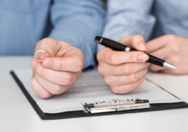 Person holding a wedding ring in one hand and a pen in the other, reviewing divorce paperwork on a clipboard.