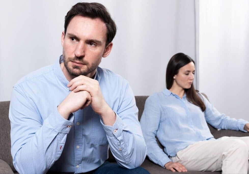 A man with short dark hair and a beard wearing a light blue button-up shirt sits pensively with his hands clasped, looking concerned. In the background, a woman with long dark hair wearing a similar blue shirt sits on the same couch but facing away from him.