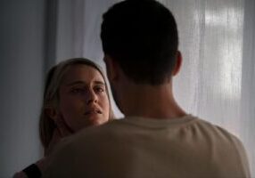 The image shows a tense scene between two people in what appears to be an indoor setting near a window. A person with blonde hair is shown facing the camera with a concerned or apprehensive expression, while another person (shown from behind wearing a light-colored shirt) stands in front of them.