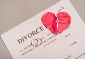 A divorce document with two wedding rings placed on it and a torn red paper heart above. The document shows "DIVORCE" in large text at the top, with legal text visible below.