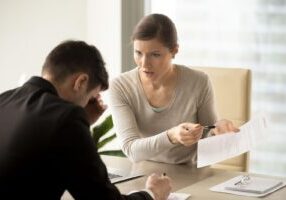 A woman in a beige sweater looking sternly at divorce paperwork while pointing at documents, sitting across from a visibly distressed man in a black suit during what appears to be a divorce consultation. A laptop and legal documents are spread across the desk between them.