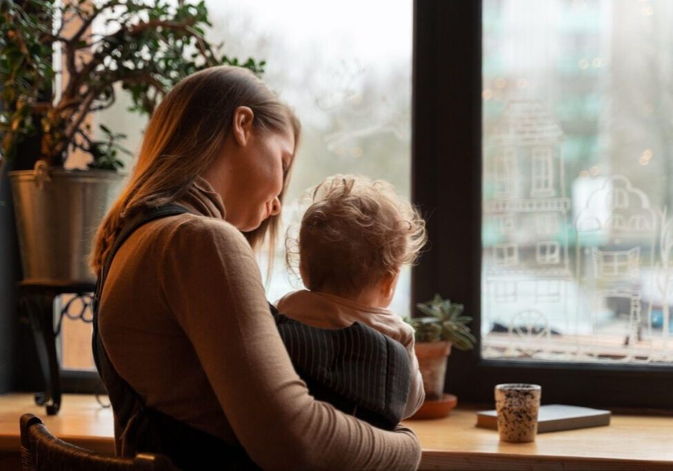 A woman with long hair wearing a light brown sweater sits by a window, holding a young child with curly hair who is wearing a dark striped outfit. They appear to be looking out at a foggy view.